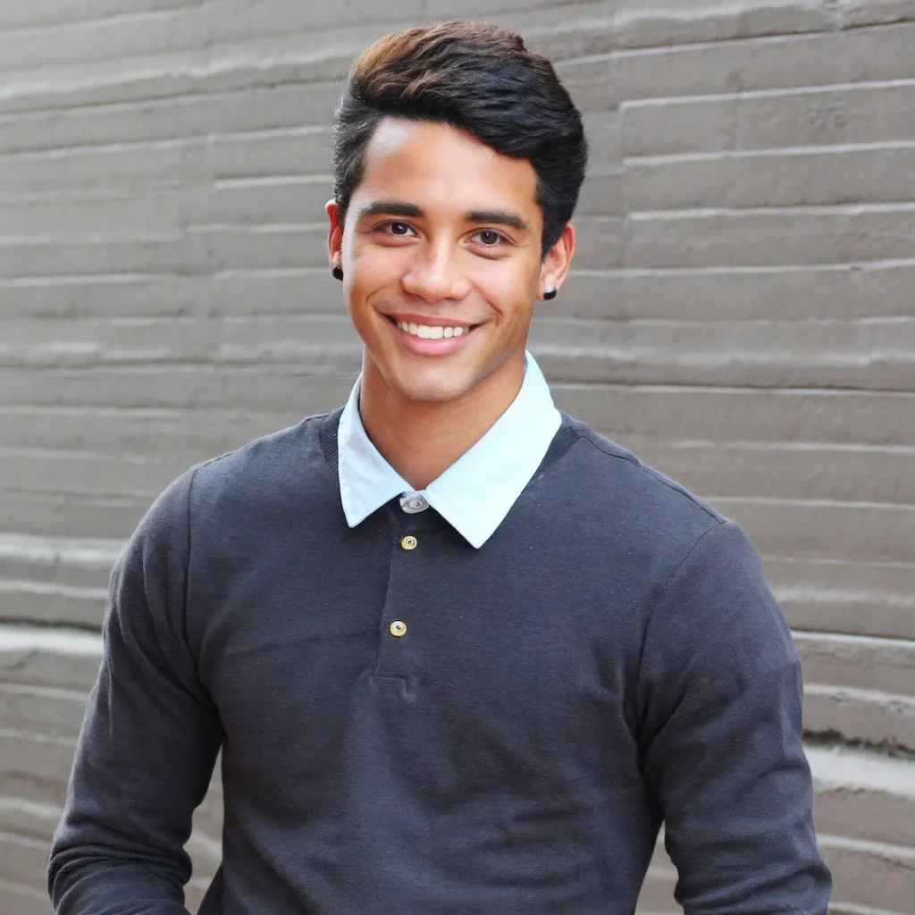 cheerful young man named alex, with neatly styled dark hair, wearing a preppy grey sweater and blue shirt, in front of a grey wall